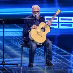 Niewidomy piosenkarz z gitarą na scenie, w okularach i z mikrofonem przed sobą, gotowy do występu.