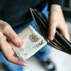 Ręce trzymają polskie banknoty 200 zł i portfel. Może to symbolizować świadczenie pielęgnacyjne lub inne wydatki.