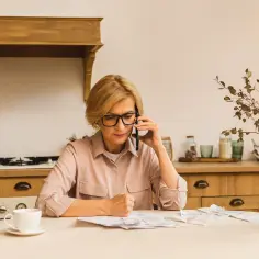 Kobieta w okularach rozmawia przez telefon, siedząc przy stole w kuchni. Na stole rachunki, filiżanka i dzbanek. Zarządzanie wspólnym gospodarstwem domowym bywa wyzwaniem.