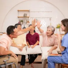 Radosna grupa wsparcia świętuje sukces. Seniorzy i opiekunka śmieją się i klaszczą, czuć pozytywną energię.