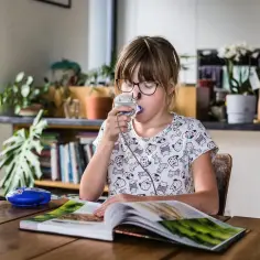 Dziewczynka w okularach używa inhalatora, czytając książkę przy stole.