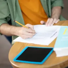 Osoba pisze ołówkiem na arkuszu odpowiedzi, obok leży książka i tablet.