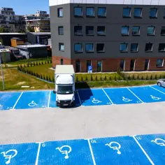 Niebieskie miejsca parkingowe dla niepełnosprawnych z białymi symbolami wózka inwalidzkiego, pod blokiem mieszkalnym.