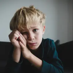Chłopiec z blond włosami opiera głowę na dłoniach, patrząc w stronę obiektywu.