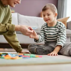 Kobieta podaje dziecku z zespołem Downa niebieską zabawkę, siedzą na dywanie wśród kolorowych klocków.