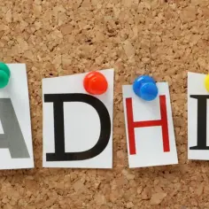 Napis ADHD z kolorowymi pinezkami na tablicy korkowej.