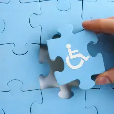 Dłoń wkłada niebieską łamigłówkę z symbolem osoby na wózku inwalidzkim w brakujące miejsce.
