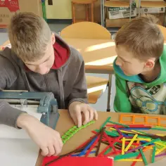 Dwóch chłopców przy stole, jeden korzysta z maszyny Braille'a, drugi bawi się klockami.