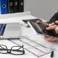 Biznesmen liczy koszty likwidacji zakładu pracy RP-7. Na biurku laptop, kalkulator, dokumenty i okulary.