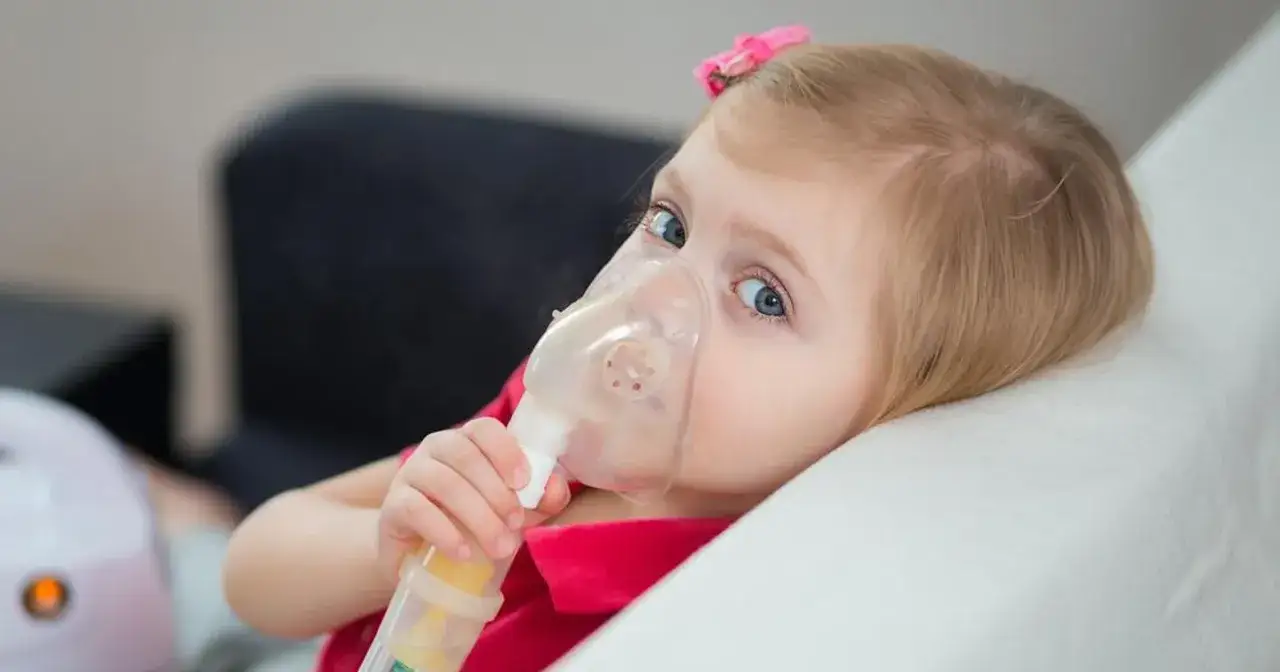 Mała dziewczynka z niebieskimi oczami używa inhalatora. To jeden z objawów mukowiscydozy, który wymaga leczenia.