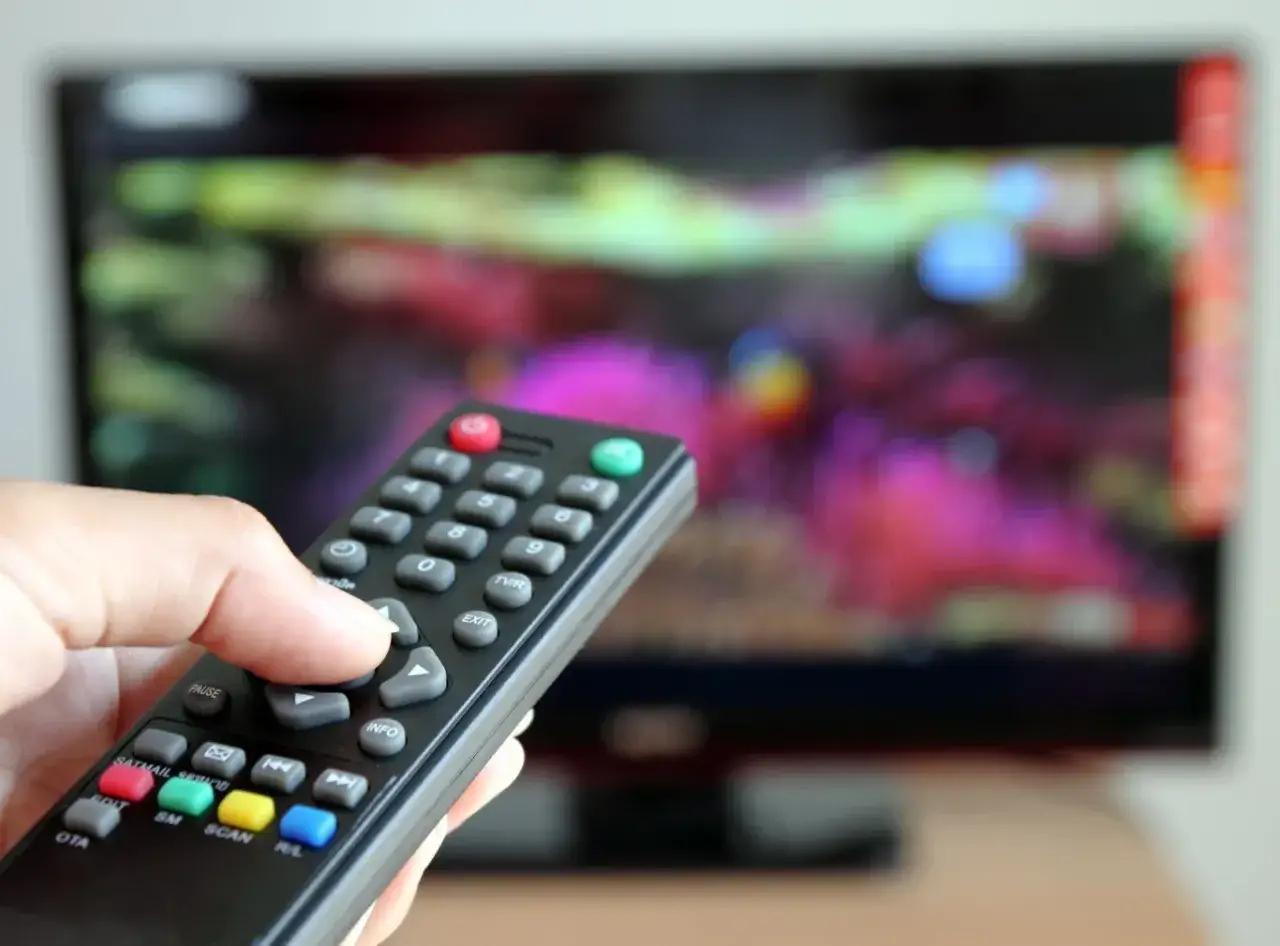 Dłoń trzyma pilot, celując w ekran telewizora. Może to symbolizować decyzję o zwolnieniu z abonamentu RTV i zmianie sposobu spędzania wolnego czasu.