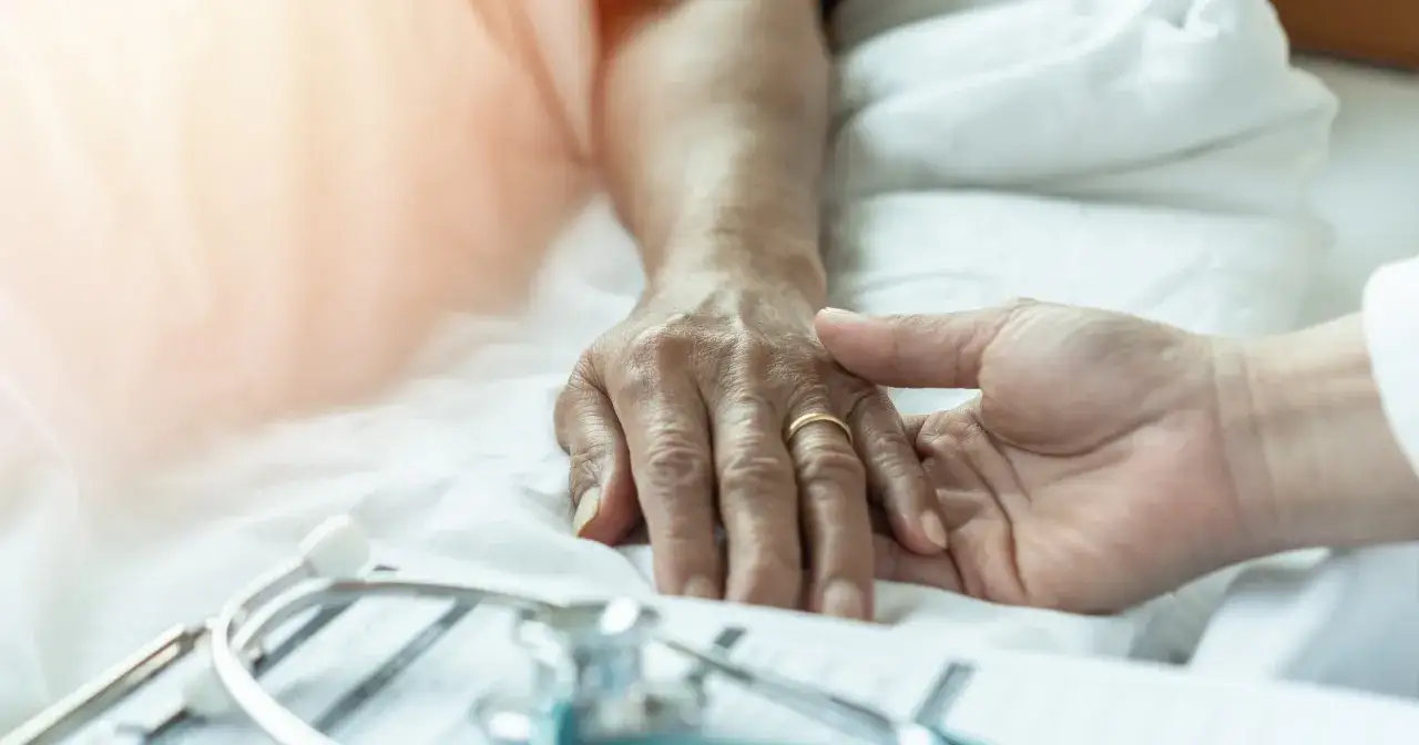 Dłoń opiekuna obejmuje dłoń starszej osoby, symbolizując troskę i wsparcie. To obraz opieki nad rodzicem zamiast pracy, pełen czułości.