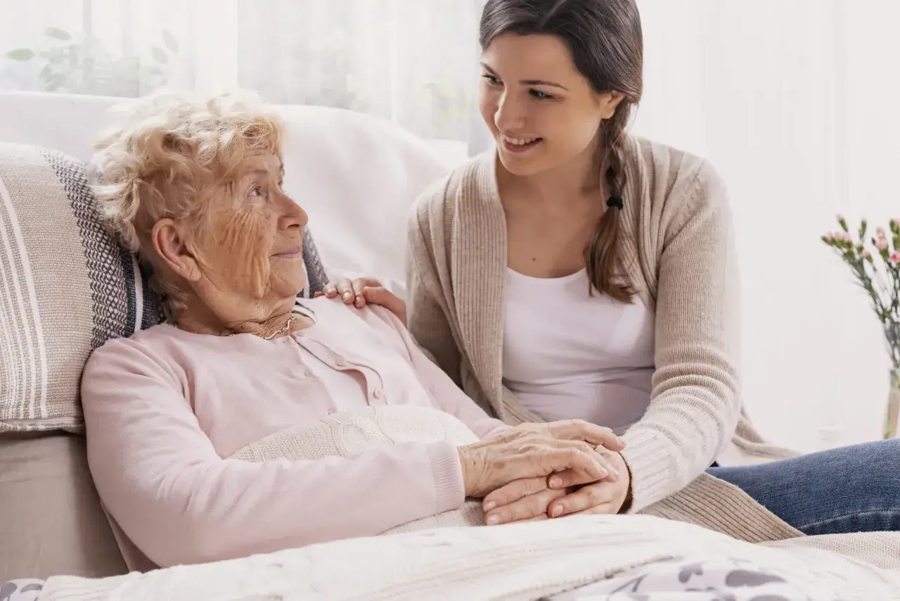 Młoda kobieta troskliwie trzyma za rękę starszą panią, która leży pod kocem. To obraz czułości i wsparcia, pokazujący, na co umierają chorzy na Alzheimera – na utratę bliskich i wspomnień.